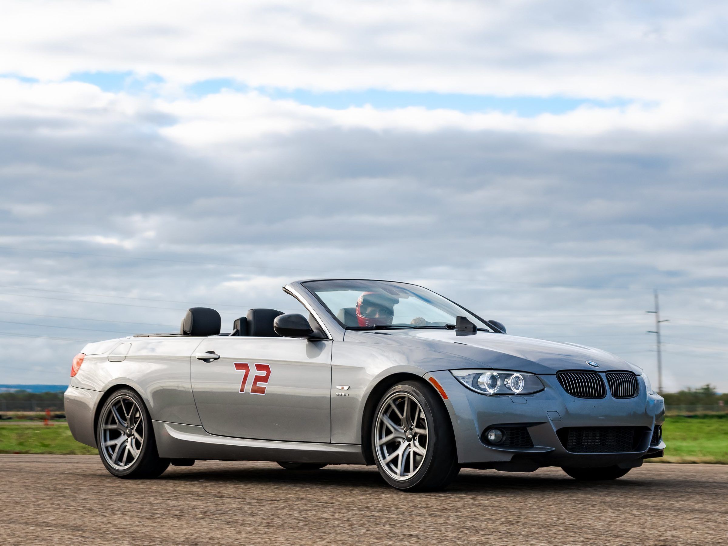 A driver in a red helmet cornering a grey convertible, with a small timing unit on the hood.