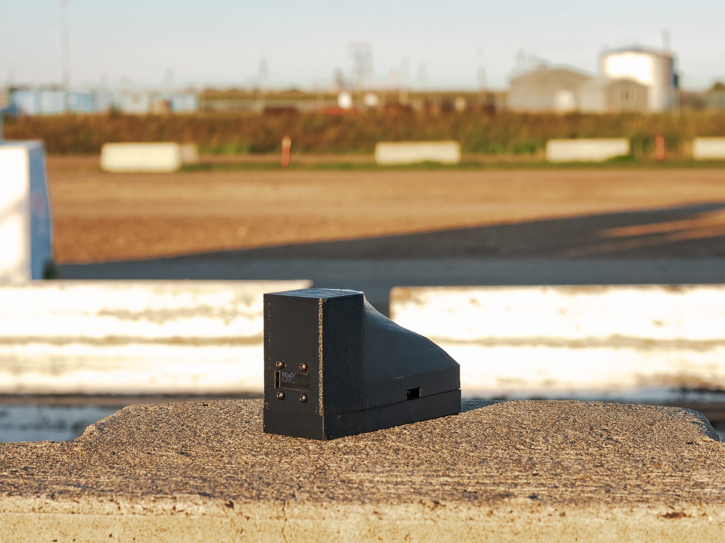 A black 3D-printed wedge device with a small display, on a trackside barrier.