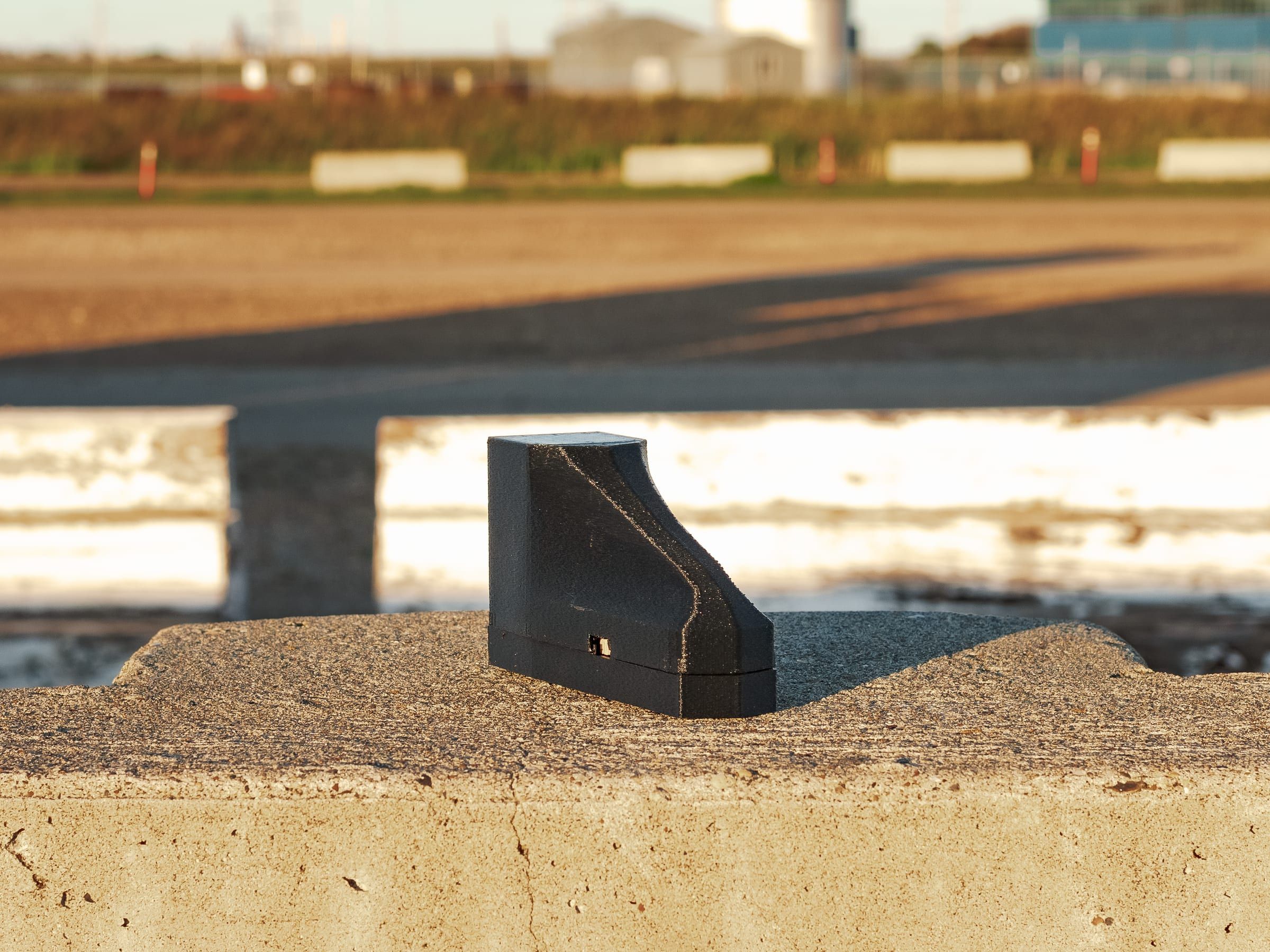 A black 3D-printed wedge device in profile, on a trackside barrier.